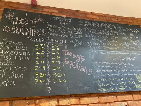 An ordinary looking menu chalkboard in a cafe with prices of drinks but there are equations and dense text in the bottom right corner.