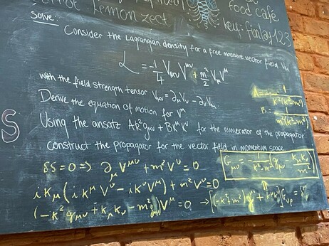 A photo showing the bottom right of the chalkboard cafe menu. A maths/physics problem is written in white chalk and there are many equations in the problem and the yellow chalk solution.
