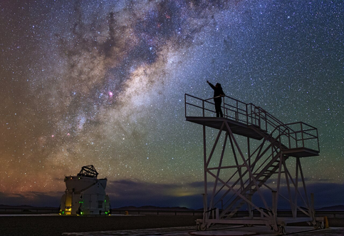 A long exposure astrophotograph showing the core of the Milky Way, with shades of orange and blue and dark lanes blocking part of the view. To the right, a person is standing on a metallic platform raised above the ground, silhouetted against the night sky and pretending to reach out to the Milky Way with an extended arm. Below the Milky Way, a small telescope is pointing up.