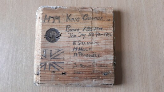 A square of wood with a coin inserted in top left corner and then pencil drawn union jack flag in bottom left and a handwritten note reading: HM King George V Buried 1.30pm this day 28 Jan 1936 E. Sellers, H.Holy, A. Pragnell, Foster & Dicksee Rugby & London.