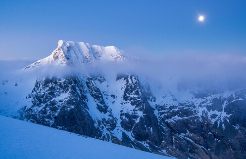Ben Nevis lit by moonlight. A snow covered mountain with a thin layer of cloud, with the moon above.