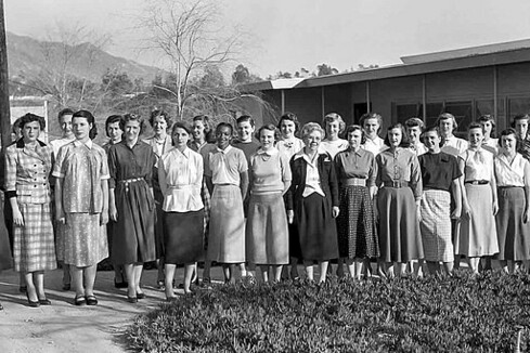 A photograph of the first 25 women to become "Human Computers" at the Jet Propulsion Laboratory. They are standing outside the lab facility in the Los Angeles area, with the hills in the background. The women are stand in two rows across. The building is in the background to the right.