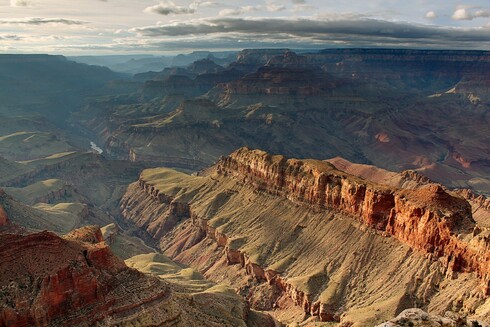 A photograph of the Grand Canyon at sunset. In the foreground, a high cliff runs from the lower right to the upper left, with orange and yellow layers on its walls. A river flows to its left. Other cliffs and valleys can be seen further behind, all the way to the horizon. Sunlight comes from the upper left.