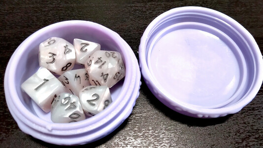 Photo: Closeup of a macaron-shaped screw-top container holding a tiny set of white, pearlescent polyhedral dice.