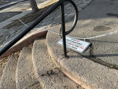 Concrete stairs with a metal railing. A sign hanging from a chain and formerly suspended over the stairs has been stomped onto the ground. It reads, “These stairs are not winter maintained”.