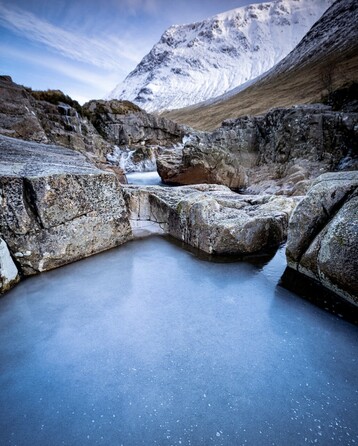 In the foreground lies a rock pool, frozen solid during winter. The cold blue ice contains very small white bubbles contrasting against the solid grey rock. Beyond, the mountain, Stob a’ Ghlais Choire, towers above the River Etive and is covered in snow and ice. Blue skies with light white cloud complete the cold scene.