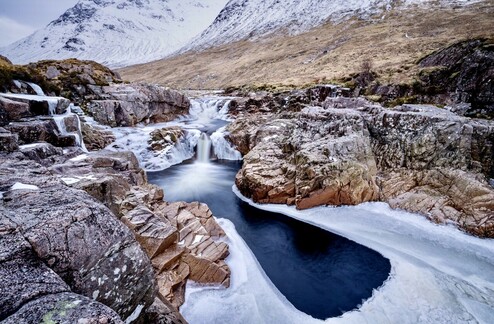 A river runs through a section of slick frozen rock in Glen Etive. Flowing down over two waterfalls, the power of the water prevents a portion of the surface from freezing over and so the water appears to run from a frozen section down into a dark blue pool before disappearing under the ice again. The river remains frozen for several miles downstream. The cold seems to permeate this image.