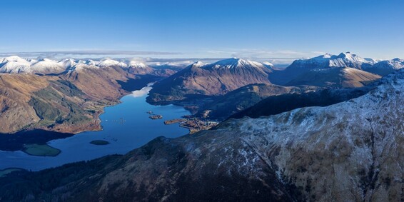 A clear blue sky day in the Glencoe area provides a panorama of the mountains in the region. The foreground is filled by the frosty ground of the ridge of mountain Sgorr Dhearg. The middle ground contains the calm still waters of Loch Leven and the village of Glencoe. In the distance, the mountains of Glen Coe and the Mamore Forest are snow-capped against a blue sky.