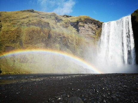 A waterfall on the right side of the photo. On the left side a cliff. In front of the cliff a rainbow (from the ground up to less than the middle of the cliff). Above the cliff the blue sky with a small white cloud.