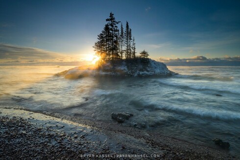 Photo of an island surrounded by waves and sea smoke. The sun is breaking through the trees on the island. The colors are cool blue except for the sun area.