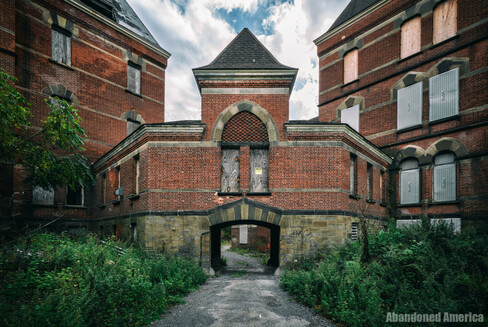 The exterior of a red brick state hospital building. Two four story buildings are on the left and right, and they are connected by a one-story brick walkway with an arched window and a walkway underneath that leads toward the camera. The windows are all boarded or have metal grates over them.