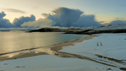 Image shows an beach covered in snow. The beach is empty except for two young children in the distance running towards the sea.there are dark clouds along the horizon and a slight warm glow of sunset