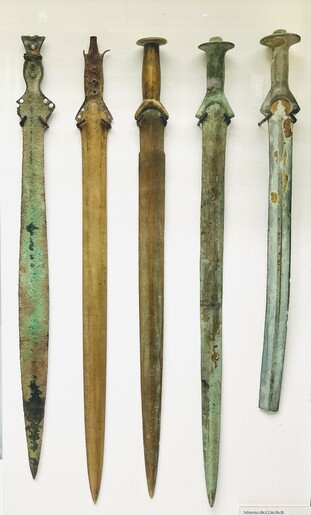 A Museum display. Five swords, some greenish, some brownish, next to each other on a white background.