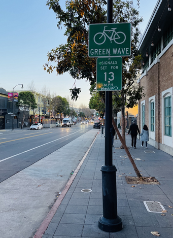 Image of a street sign at the side of the road that reads green wave and that says that traffic signals are timed for 13 miles per hour