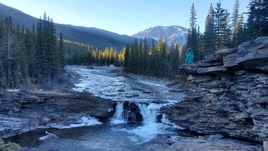 A woman sits on a rock ledge overlooking a small waterfall along a river that is just starting to ice over at the edges, with pine trees and a snow-dusted mountain peak in the background