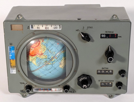 The Globus INK-2S is a grayish metal box with a colorful rotating globe in the middle under a plastic dome. The box has six knobs, three indicators with rotating digits, and three dial-like indicators. The knobs and displays are labeled with Russian text.