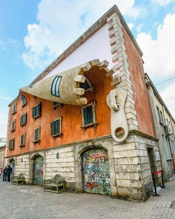 A three story stucco building in Italy showing an art installation featuring an oversized zipper running up the side of the building. The zipper is open and the stucco wall rolls open like fabric.
