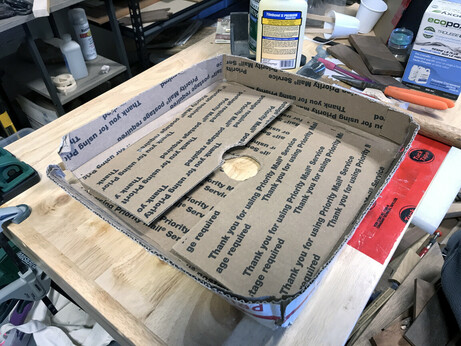 Part of a repurposed cardboard box, with a hole cut in the middle to feed a vacuum hose through, sitting on a work bench (surrounded by too much stuff I need to put away).