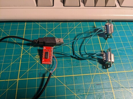 Small red PCB board with MAX232 chip, there are two DB 9 plugs and a USB A cable connected to it.