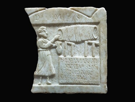 The fragment of a Roman relief shows a woman with a wine jug in front of a barrel. Below the barrel is an inscription giving the name of the deceased, her age, and her husband.