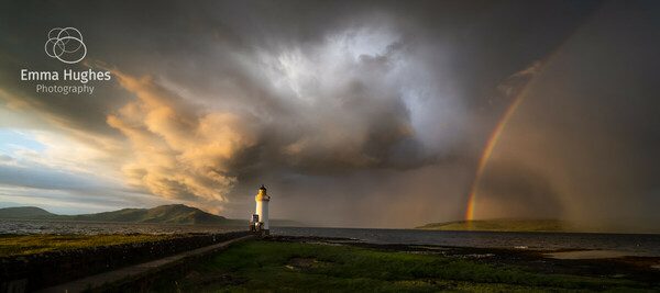 landscape view of a white lighthouse, on the right hand side there is a rainbow curving out of shot in front of dark clouds, whose rainfall obscures the horizon, moving over the lighthouse the sky and clouds explode out catching the sunlight that shines on the lighthouse. The sky on the left lightens and reveals a pale blue sky as it stretches away from the small dark violent squall