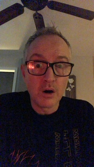 this is a late-night selfie of me — jamie! i'm a YOUTHFUL 50yo white male; i'm wearing black glasses and a black crew-necked shirt (i believe people might be starting to realize that this is a recurring theme). i'm sitting in my recording studio ... lately i'm always sitting in my recording studio! this makes sense, because i'm a record producer, but a change of scenery would be nice. maybe tomorrow we'll take a walk or something. i always say that. but i have some good time right now to work on this record i'm mixing, so i'm making hay. anyway, the picture! it's grainy and low-light, but it looks like a nice environment ... because it is. there's a dark brown ceiling fan with five blades, some homemade art on the wall that someone made based on an older album of ours,and that's about all you can see. really it's just a selfie of a tired and vaguely stunned man.