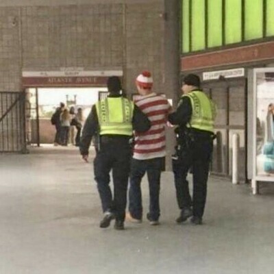 Man in Where’s Waldo costume being detained by 2 members of law enforcement.