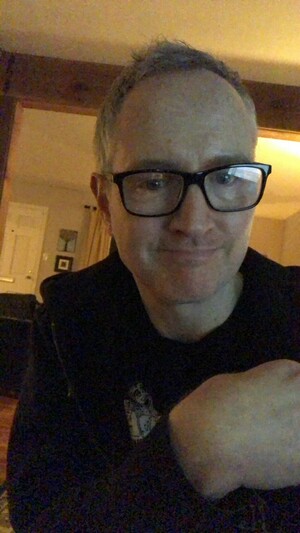 this is a grainy nighttime selfie of me, jamie hill. i'm a white male; per usual, i'm wearing black glasses, a black tee shirt, and a black hoodie. for the record, i'm also wearing black pants and socks. i'm in my studio, although i'm facing all the fancy gear, so you just see a couple of rooms behind me. there's some art on the wall; the transition between the two rooms is trimmed in wide raw cedar boards. i'm smiling!