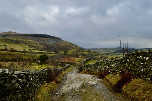 An overcast sky, muted colours. A lane with old stone walls leading towards a craggy hill. The floor of the lane is partly a solid sheet of rock on a rough slant.