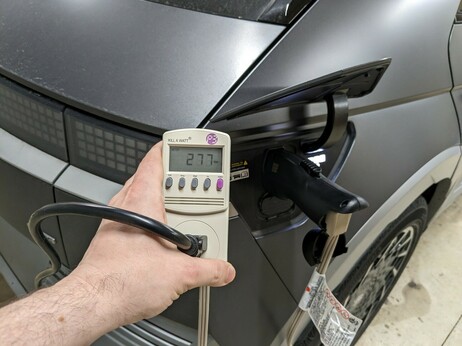 My left hand holding up a kilowatt power meter showing 277 watts of consumption, the meter is next to an adapter plugged into the charge port of a Hyundai Ioniq 5