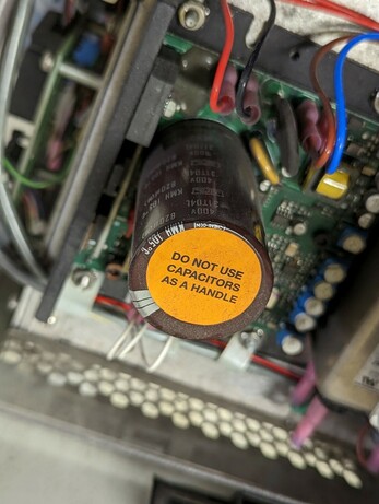 Photo of a circuit board with a large electrolytic capacitor. The capacitor has a bright orange label saying "Do not use capacitors as a handle"
