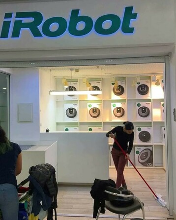 The store front of a small iRobot shop, possibly in a mall. Many vacuum robots of their brand are prominently displayed on the back wall. In front of them, a woman is cleaning the floor with a broom.