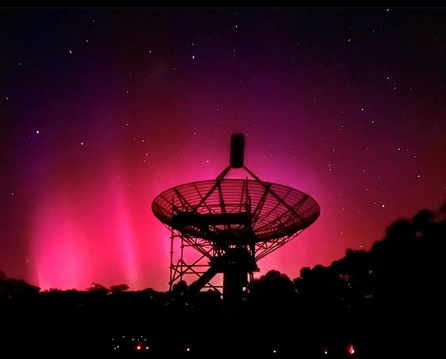 Radio telescope pointing upwards with pink aurora behind it.