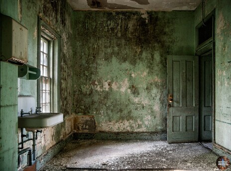 An empty room with an open doorway and a sink attached to the wall. The paint is green but age had blackened it in spots
