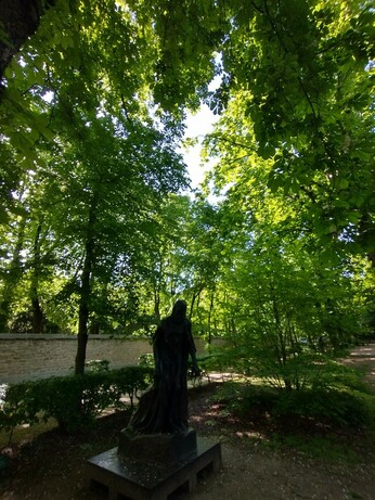 Green trees and dappled sunlight arc above a dark statue in the Rodin garden.
