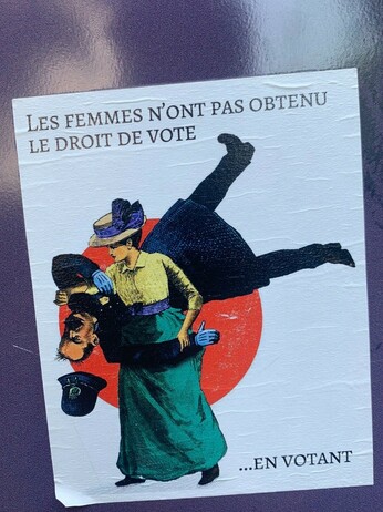 Sticker on a wall of a victorian comic of a woman throwing a cop and the words “Women didn’t get the right to vote by voting.” in French