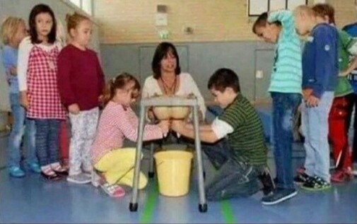 Children learning how to milk a cow from a simple table with a rubber udder underneath that releases milk into a bucket. The white teacher, wearing a cream blouse, is crouched behind the table so that it looks like her breasts are the udder