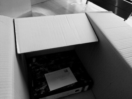 Black-and-white photo of a cardboard box containing a folder and an envelope, taken at #UniKrems.