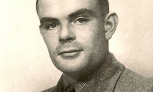 A black and white photo of Alan Turing. He is looking at the camera and the top of his head is not visible in this closely cropped image. He has dark eyes and bold eyebrows. Turing's hair is very short, which accentuates his ears. He is wearing what appears to be a gray flannel jacket, with what might be a plaid shirt.