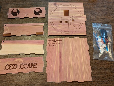 Pieces of wood, which are the sides of a box, laying flat on a table, with a small packet of electronics, including LEDs, wires and a battery holder next to it.