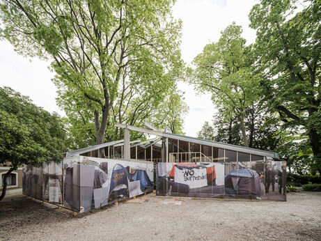 A modernist pavilion in the middle of a grove of trees. A fence of posters wrap around the structure that display scenes from a tent city.