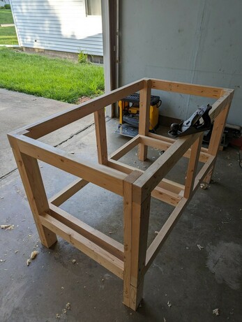 A rectangular table skeleton made with 2x4 lumber with double cross braces on the sides and middle on the bottom, the top is open. The table is in a garage with door open, looking out.