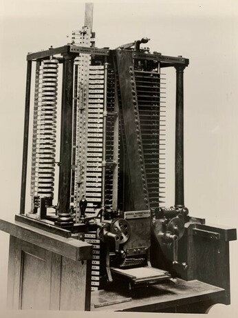 Black and white portrait orientation rectangular photograph. Designed in the shape of a lollipop; the stick (standing out at the top of the image) contained the store (memory), where the numbers were kept, and the candy held the mill (CPU), where the numbers were operated upon. It is fifteen feet tall about as big and heavy as a small locomotive.  This piece is placed on a wooden base.