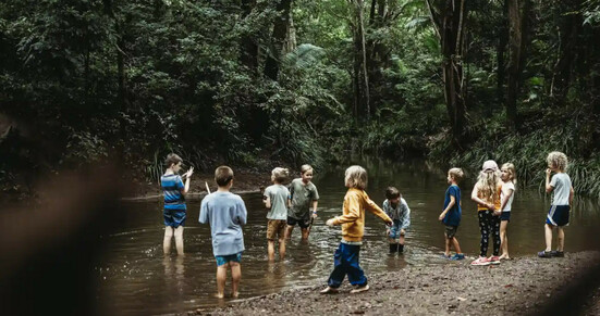 A group of children at their bush kinder program in Queensland. They paddle in a shallow pool of water surrounded by thick green forest. Photograph: Alix Clark Photography