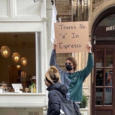 Outside of a coffee shop, A man wearing a mask holds up a cardboard sign that reads "There is no 'X' in espresso."