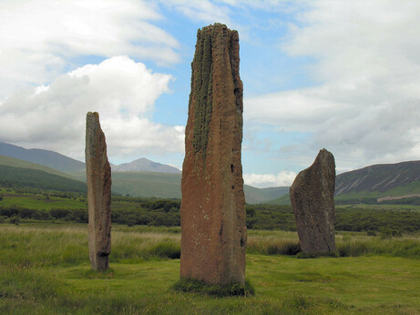 Machrie Moor. The image shows an open moorland view with hills and mountains in the background. There are three tall red standing stones in the foreground, set in a triangle with the tallest one closest to us. The sky is cloudy with blue patches.