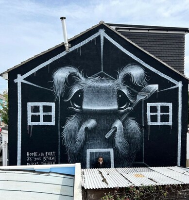 Streetartwall. On the outside of a multi-storey house is theMural of a fluffy fantasy figure been sprayed / painted. A small gray and black fluffy creature with big eyes, curved little ears and a wide nose stands there with a brush in his hand. It looks like a mixture of squirrel, mouse and rabbit. Around him is painted a house, as in a child's drawing. Title:
"Home is the port as your storm passes through, but remember your home can be found in you."
(In the photo the artist is standing in front of her mural.)