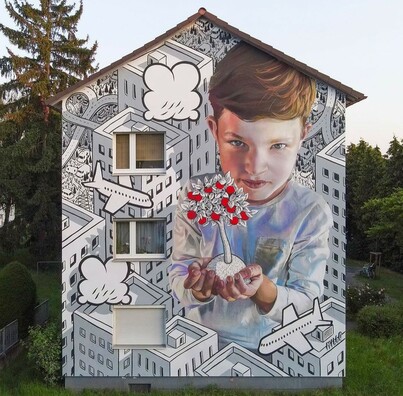 Streetartwall. On the outer wall of a two-story house was sprayed a large mural depicting a child holding an apple tree in his hands. In the background you can see a row of minimalist skyscrapers with planes and clouds flying through them. In the middle of this city, a small brown-haired boy stands holding a white tree with red fruits in his hands.
Info: Two great street artists created this mural. Millo (aka Francesco Camillo Giorgino) paired his minimalist cityscapes with Case Maclaim, known for his fantastical and realistic scenery. Both have referred to the countryside, known for its mineral springs and orchards.