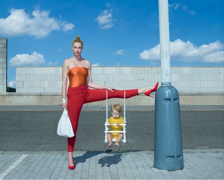 Photography. A color photo showing a woman balancing her child on one leg. The background is a blue sky with flat gray buildings and a street. A young woman wears her red hair in a knot on her head and is dressed in red stretch pants and an orange top. She wears red high-heeled shoes and stands on one leg while holding a white plastic bag. Her left leg is stretched out bolt upright and her foot rests on the gray column of a streetlight. Hanging from her leg is a child's swing with a red-haired baby sitting in it. She looks at us. Title: "The three of us"