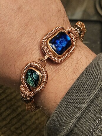 My wrist with a copper wirewrap bracelet with a green and a blue square glass gemstone on the ends, the glass has these cool trippy patterns on the inside